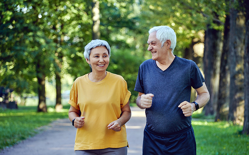 Mature smiling grey-haired fit couple jogging in the summer park. Two older joggers enjoy morning active cardio outdoors, keep healthy active lifestyle. Weight Loss, sport concept
